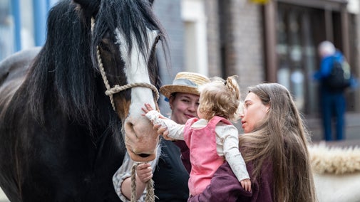 A young girl in a pink top with curly blonde hair is sat in her mother's arms, while she carefully pats a dark brown shire horse on the nose. The horse has a white streak on its nose and its looked after by a lady dressed up in Victorian clothes.
